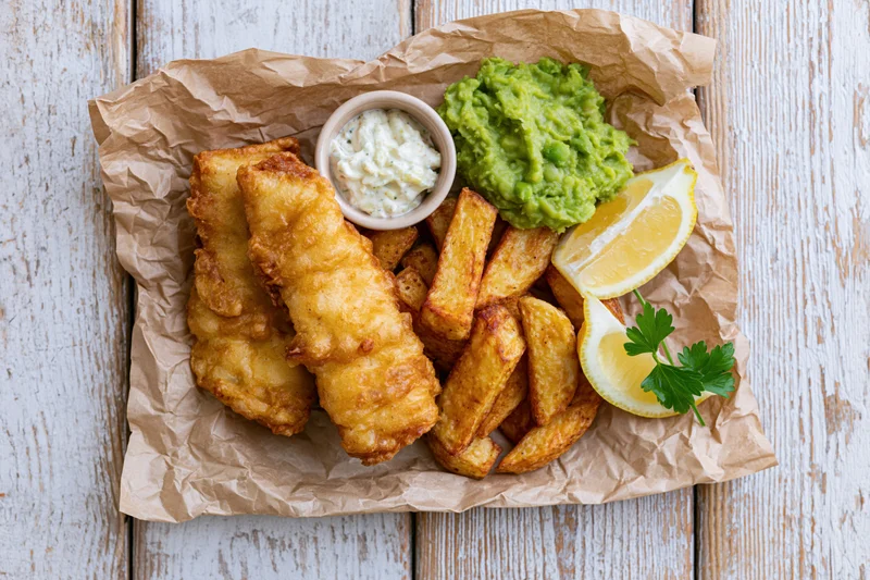 Traditional british fish and chips served on crinkled brown paper with tartar sauce, lemon, and parsley on a rustic white wooden table