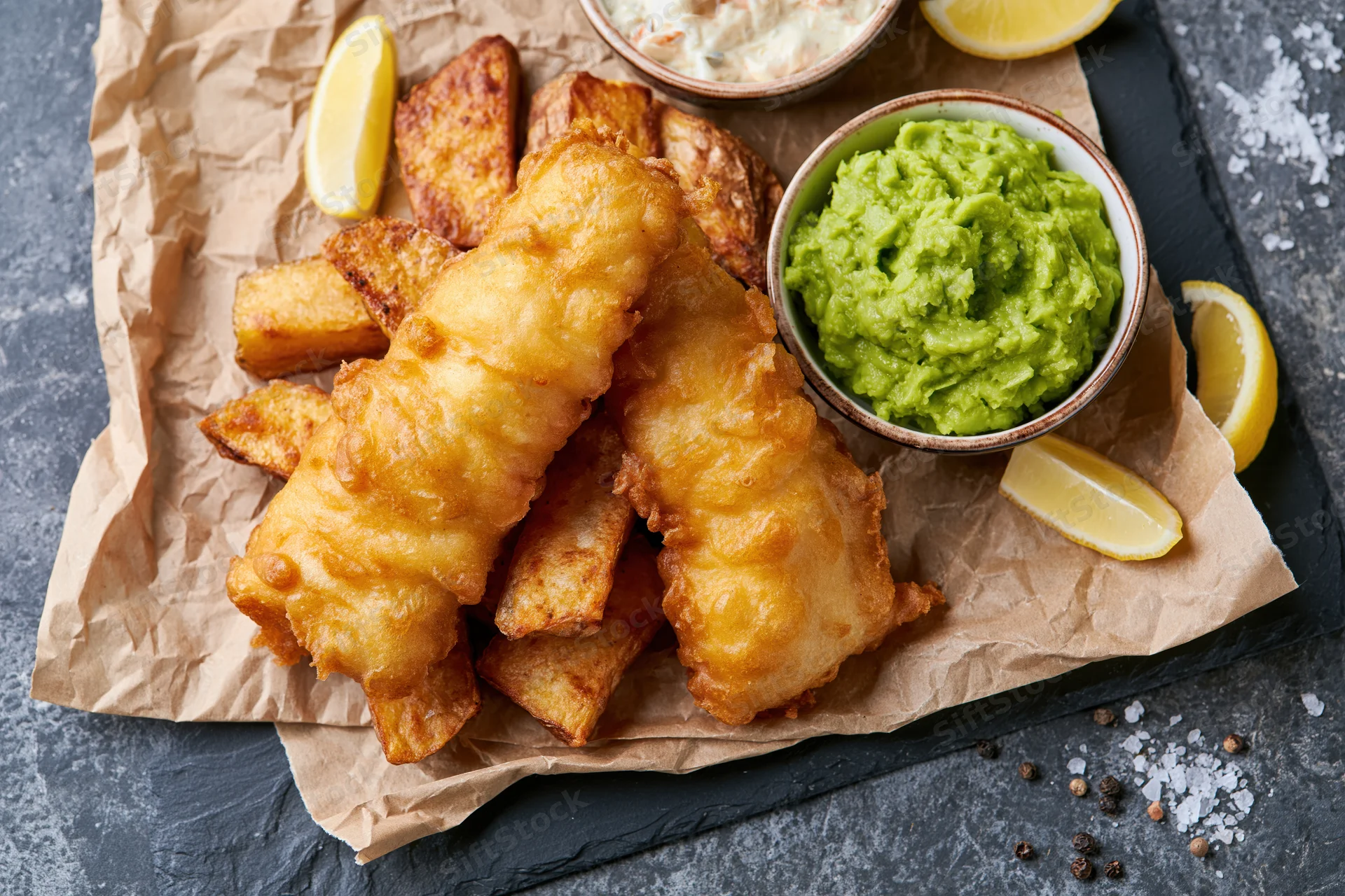 Golden battered fish and thick-cut chips served on crumpled brown paper with lemon wedges and side sauces on a slate tray
