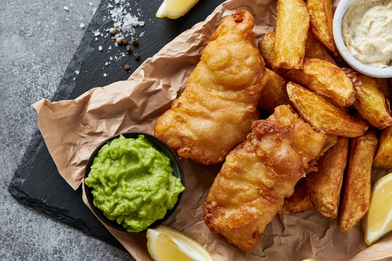 Golden fried fish and thick-cut chips served on crumpled brown paper with mushy peas and tartar sauce on a slate board