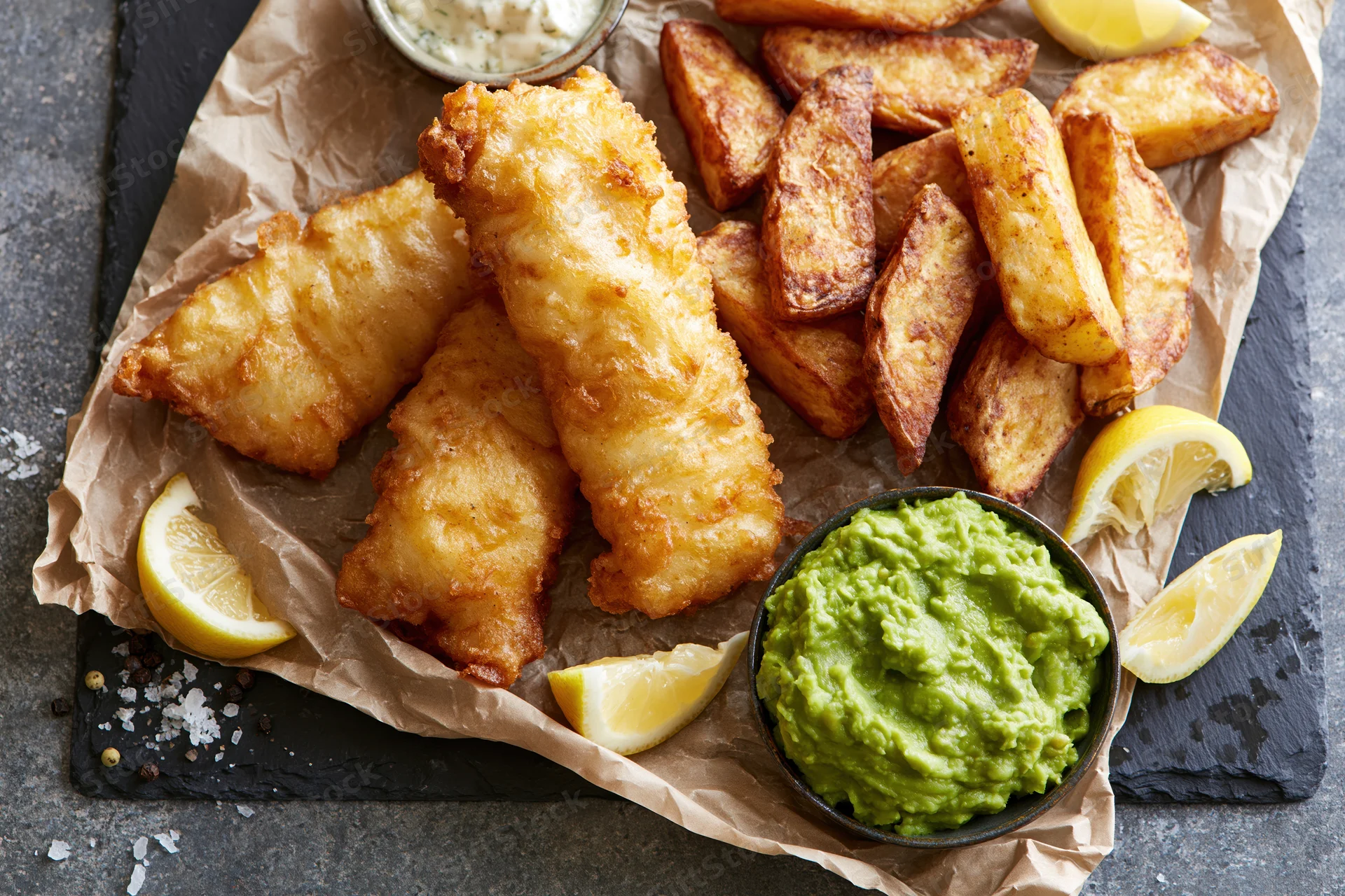 Fried fish and chips with lemon and guacamole on crumpled paper over slate plate