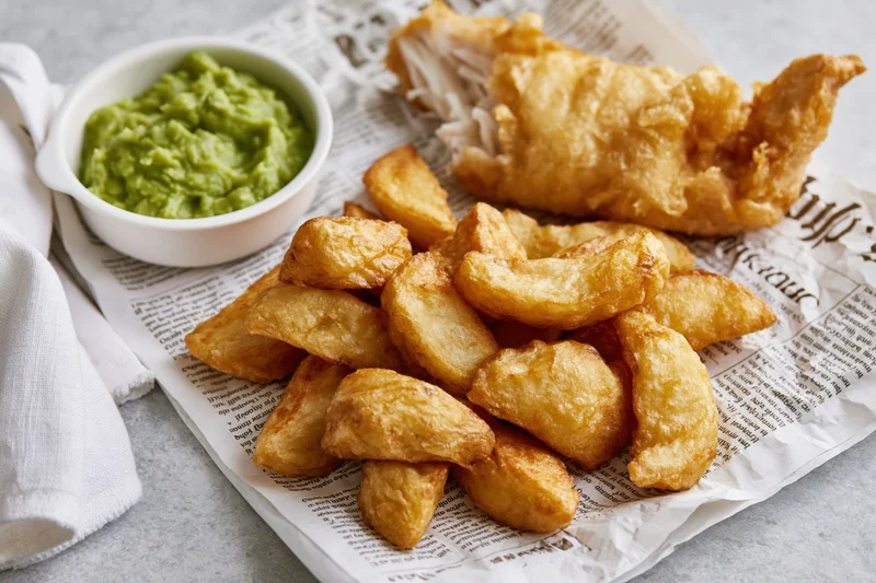 Golden fried fish and crispy chips served on printed newspaper with green guacamole in a white bowl