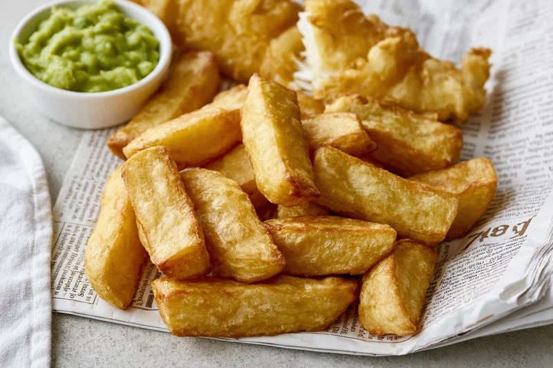 Golden fried fish and thick-cut chips served on printed newspaper with a side of mushy peas in a white bowl