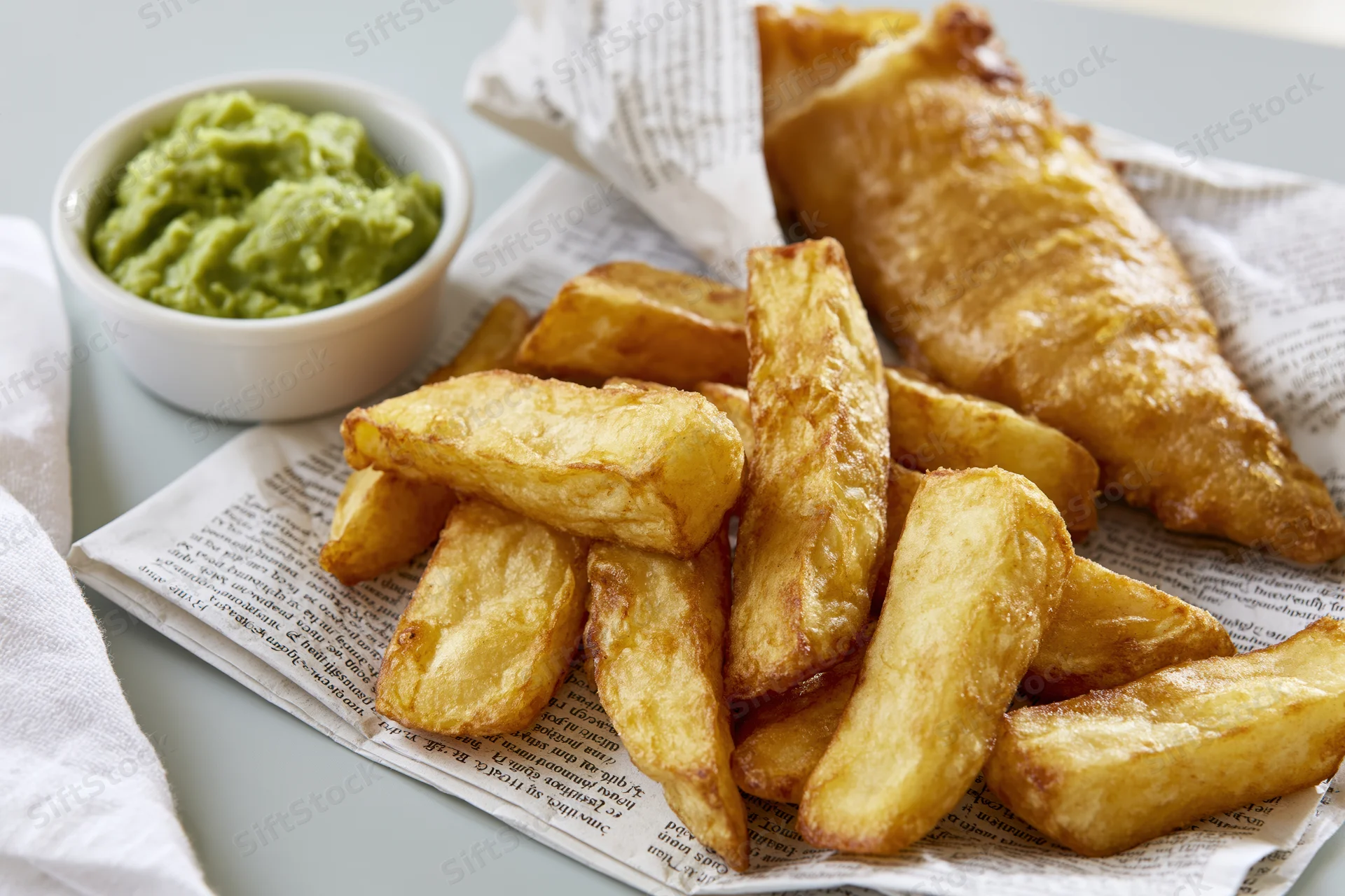 Golden fried fish and thick-cut chips served on printed newspaper with a side of mushy peas in a white bowl