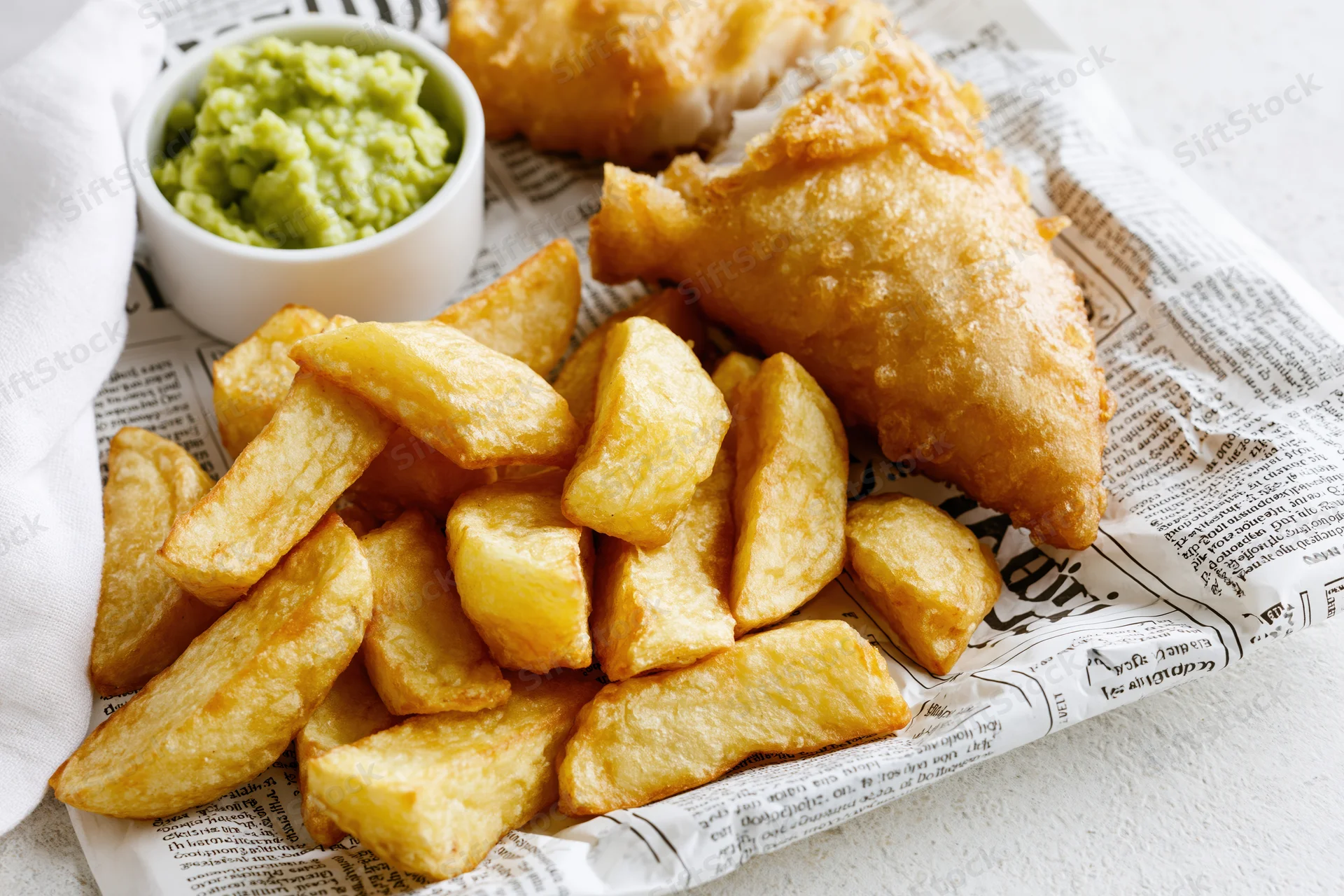 Golden fried fish and thick-cut chips served on printed newspaper with mushy peas in a white bowl