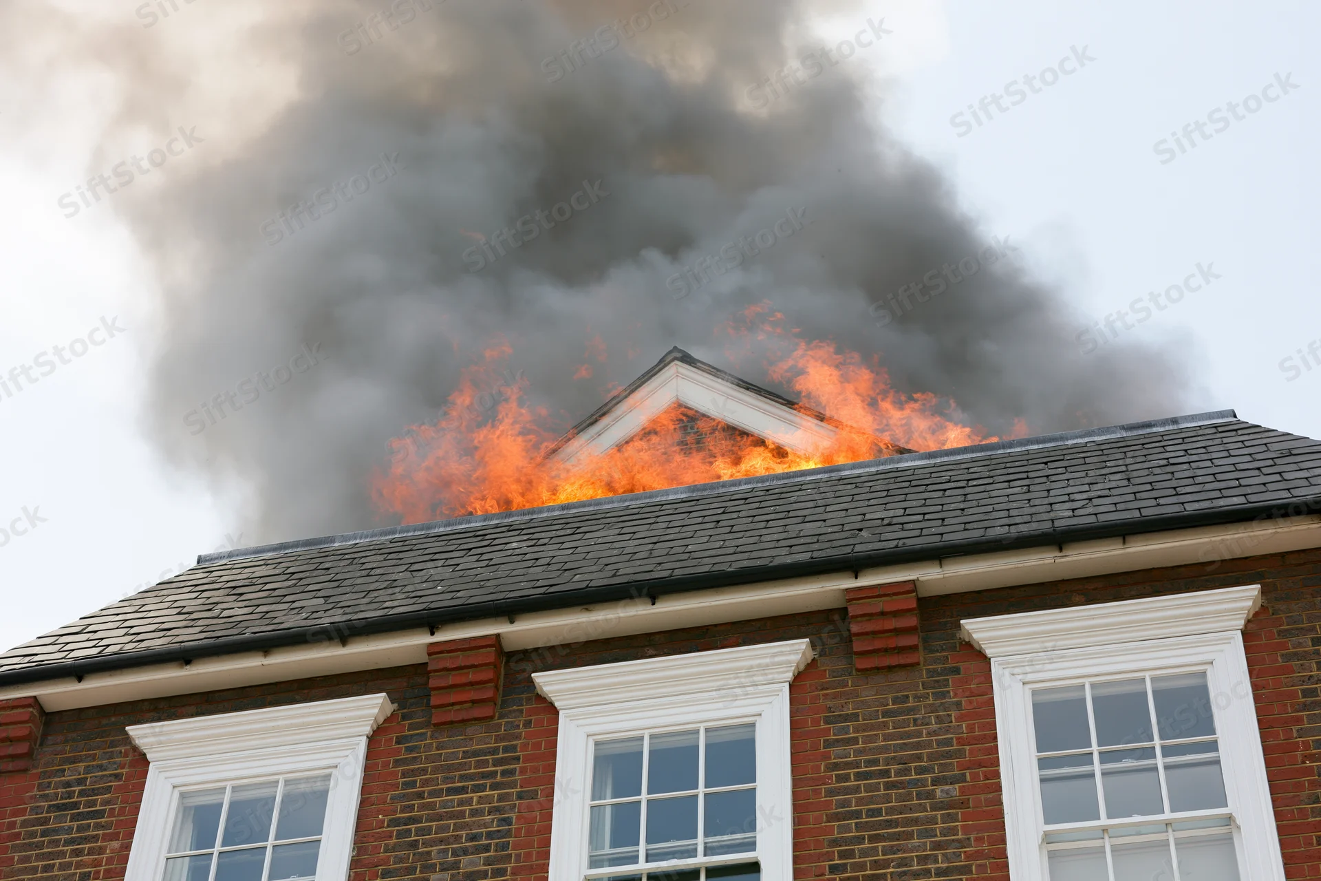 Flames engulfing the roof of a two-story brick house with smoke rising from the attic peak