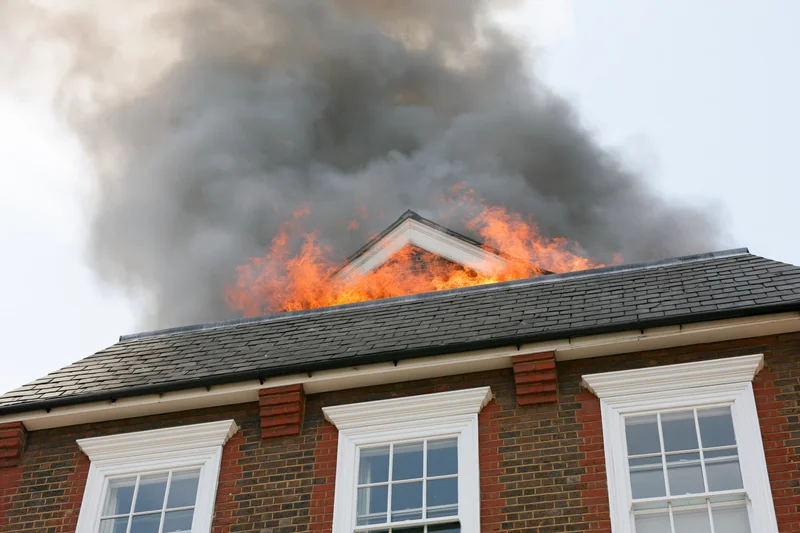 Flames engulfing the roof of a two-story brick house with smoke rising from the attic peak