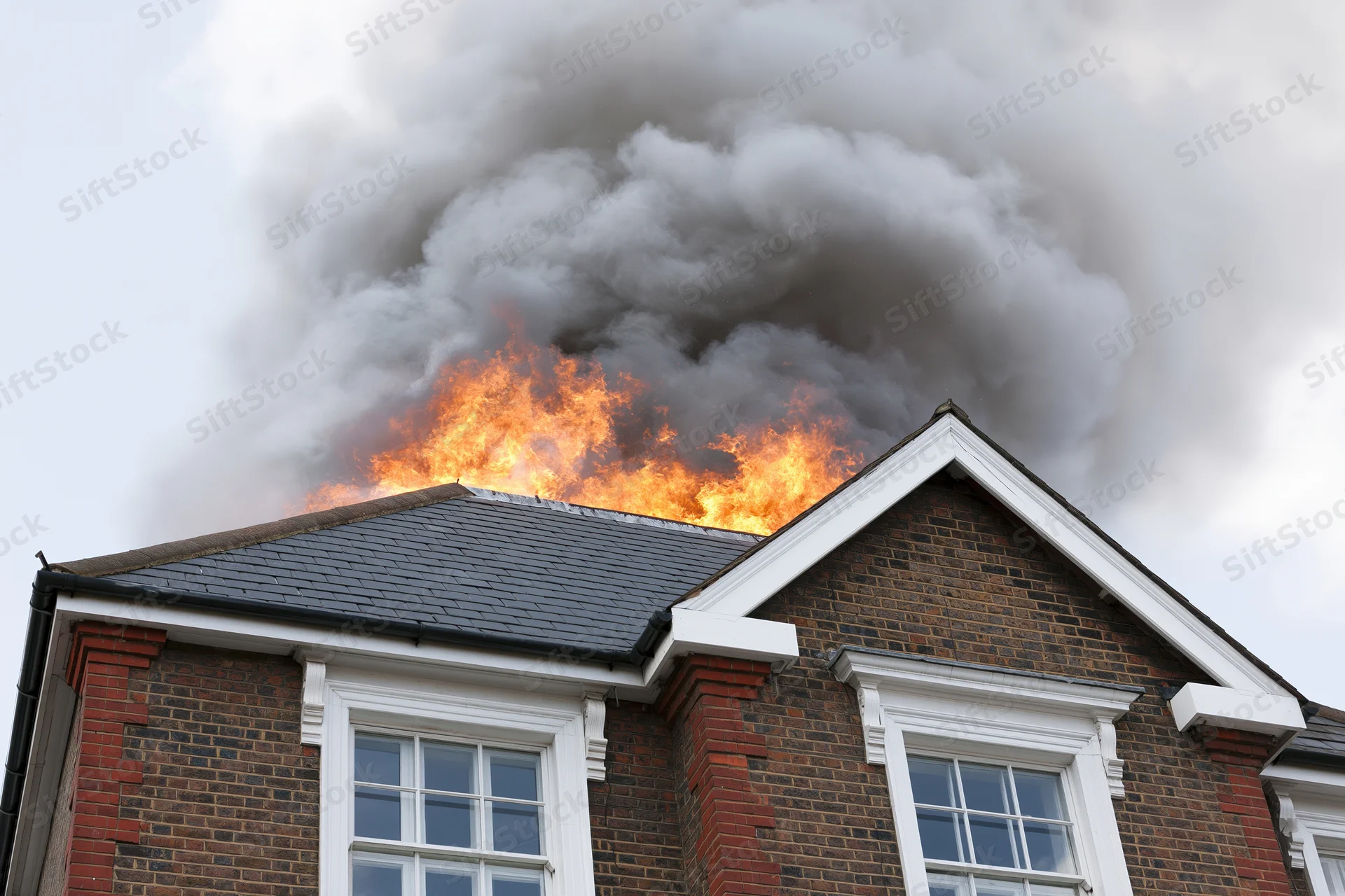 Flames erupting from the roof of a two-story brick house with thick smoke billowing upward