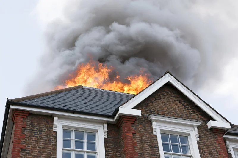 Flames erupting from the roof of a two-story brick house with thick smoke billowing upward