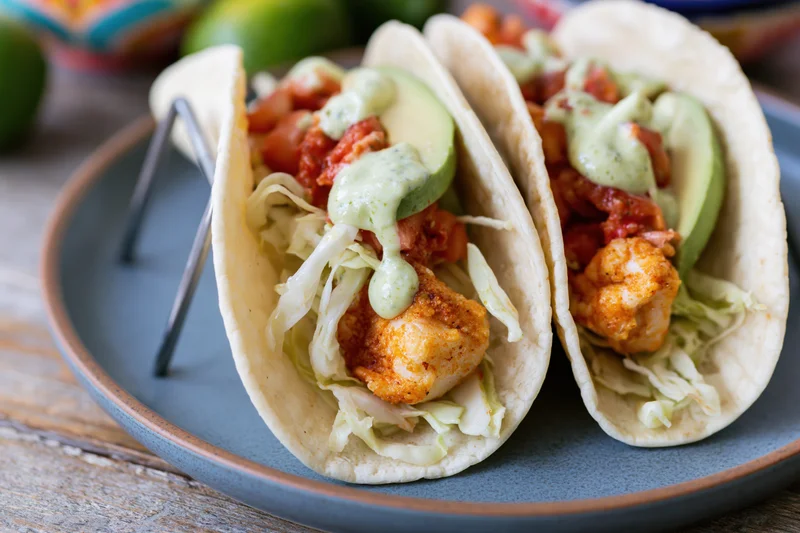 Two fish tacos in soft flour tortillas filled with cabbage avocado sauce and fried fish on a blue plate