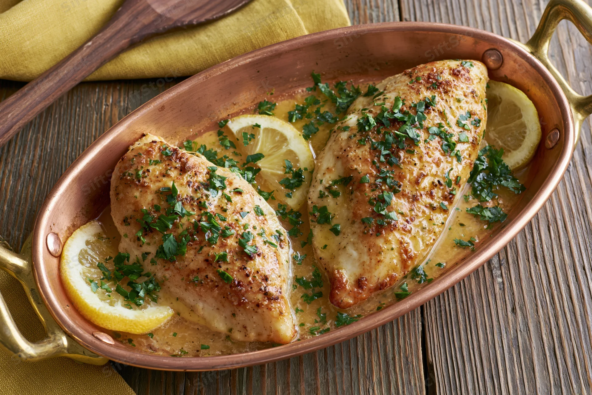 Two roasted chicken breasts in a copper oval roasting pan with lemon slices and parsley garnish on a wooden table