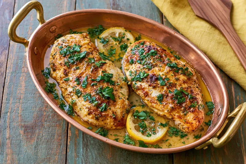 Two roasted chicken breasts in a copper oval roasting pan with lemon slices and parsley garnish on a wooden table