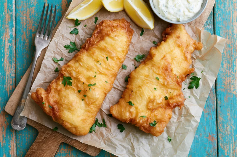 Two golden battered fish fillets on parchment paper with lemon wedges and tartar sauce on a wooden cutting board