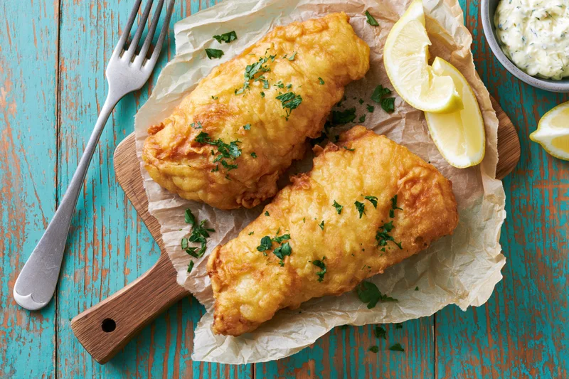Two golden battered fish fillets on parchment paper with lemon wedges and tartar sauce on a wooden cutting board