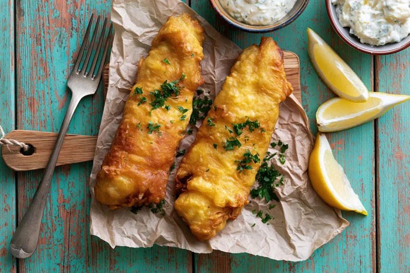 Two golden battered fish fillets on crumpled parchment paper with lemon wedges and tartar sauce on a rustic turquoise wooden table