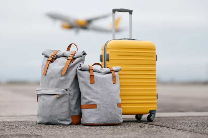 Two grey canvas backpacks with tan leather accents and a yellow rolling suitcase on tarmac with airplane in background