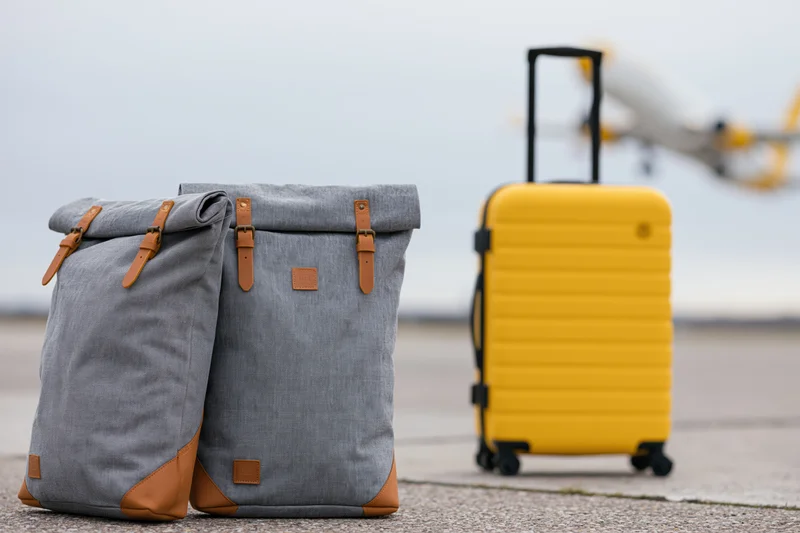 Two grey canvas backpacks with tan leather accents and a yellow suitcase on tarmac with airplane in background