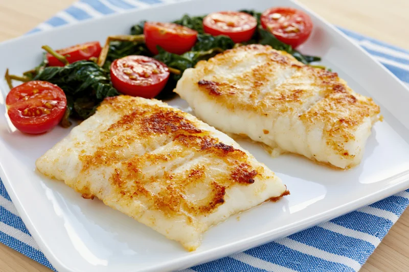 Two pan-seared white fish fillets with golden-brown crust served on a white plate with spinach and cherry tomatoes, placed on a blue and white striped napkin on a wooden table