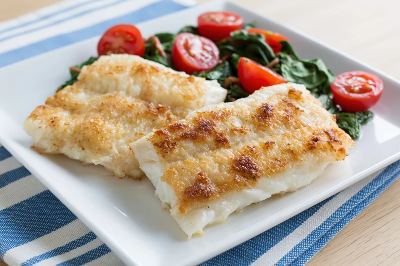 Two pan-seared white fish fillets with golden-brown crust served on a white square plate with spinach and cherry tomato salad on a blue and white striped napkin