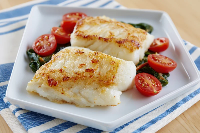 Two seared white fish fillets with cherry tomatoes and spinach on a square white plate