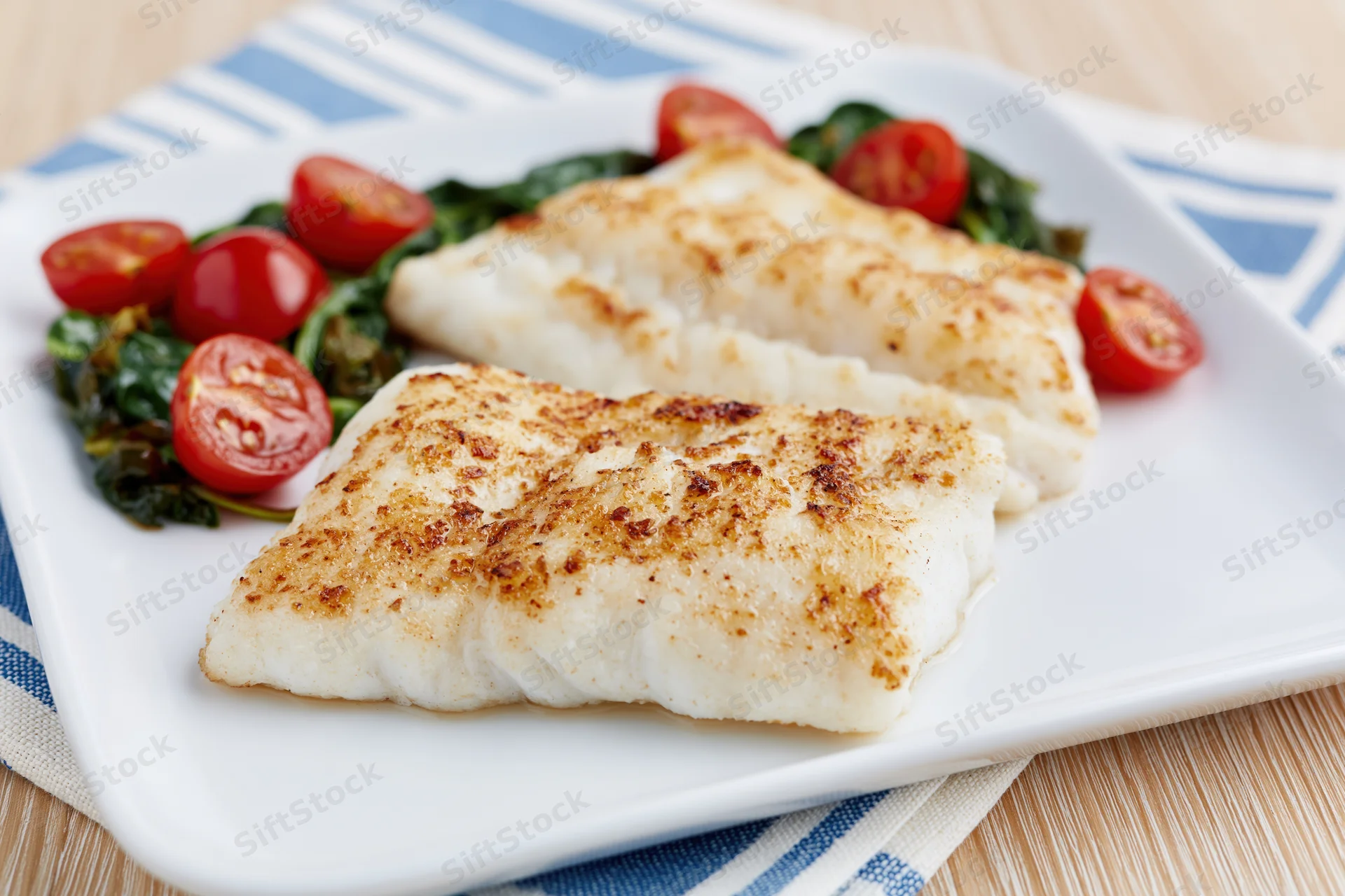 Two pan-seared white fish fillets with golden-brown crust served on a white plate with spinach and cherry tomatoes