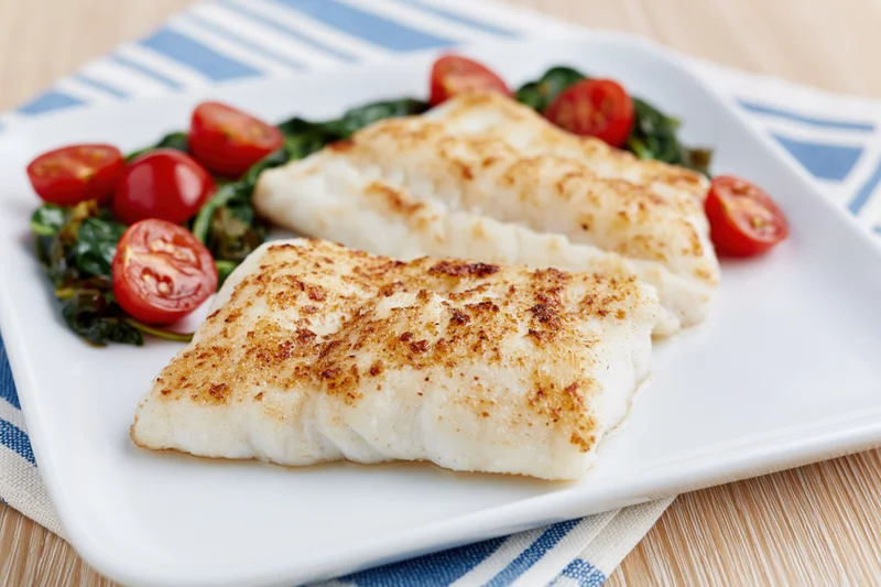 Two pan-seared white fish fillets with golden-brown crust served on a white plate with spinach and cherry tomatoes