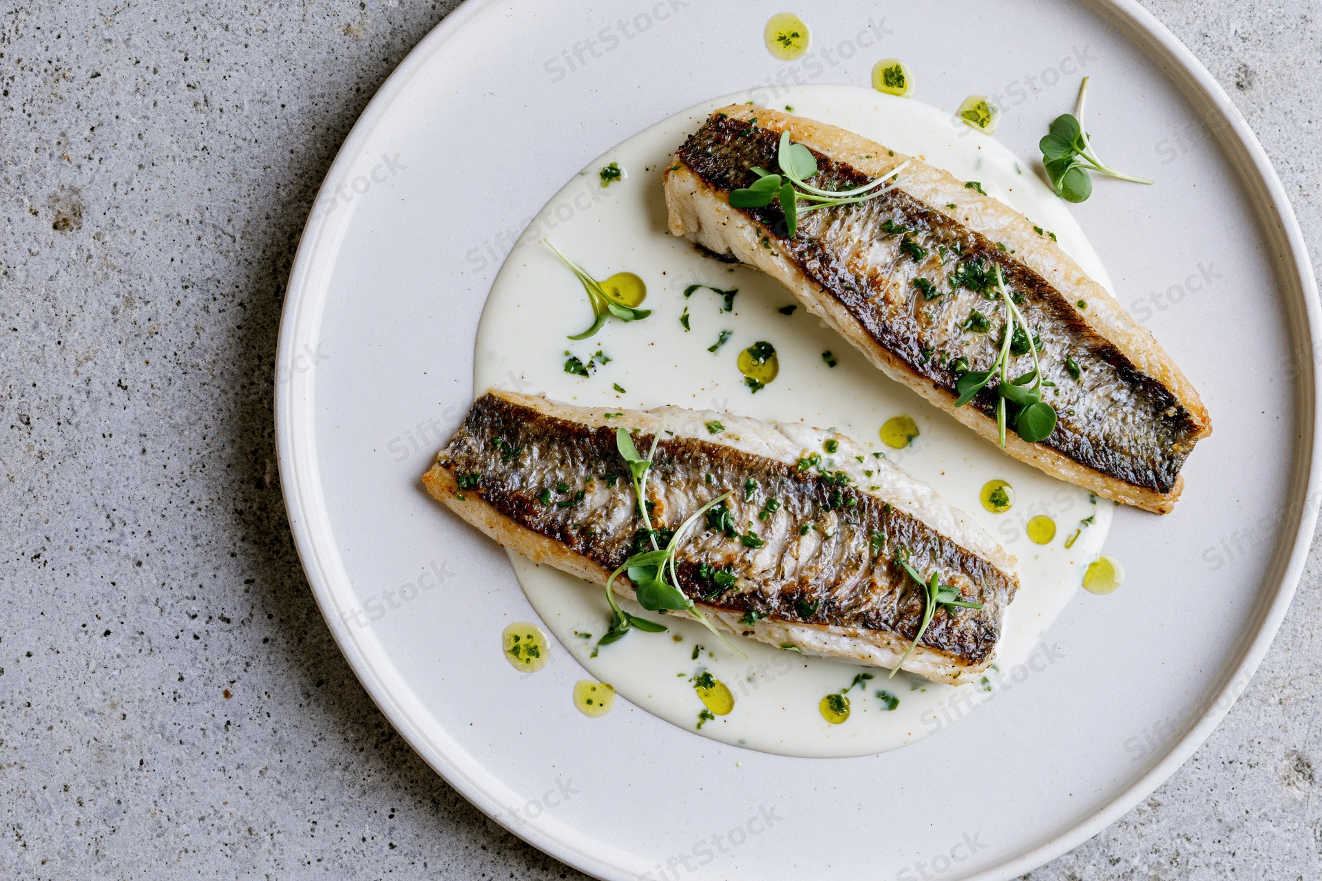 Two seared white fish fillets with golden-brown skin on a white plate with cream sauce and microgreens garnish