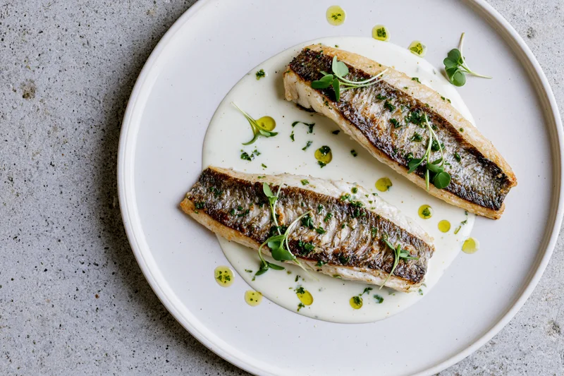 Two seared white fish fillets with golden-brown skin on a white plate with cream sauce and microgreens garnish