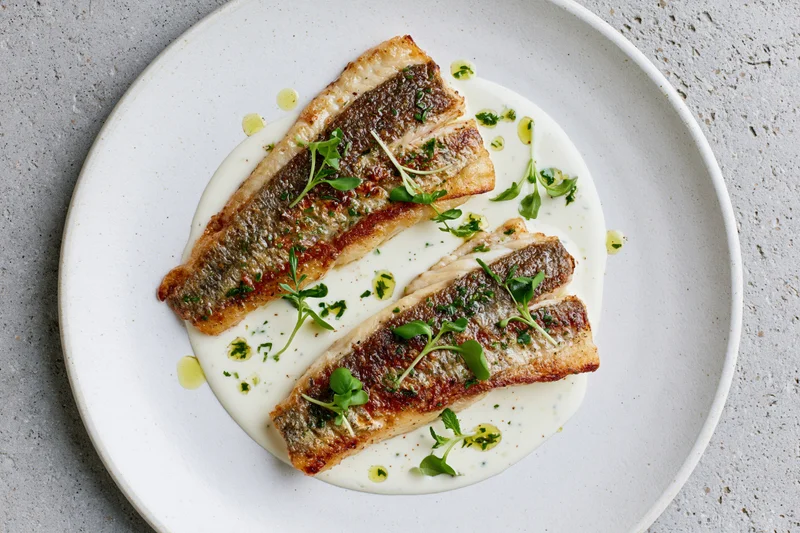Two pan-seared white fish fillets with golden-brown skin on a white plate with cream sauce and microgreens