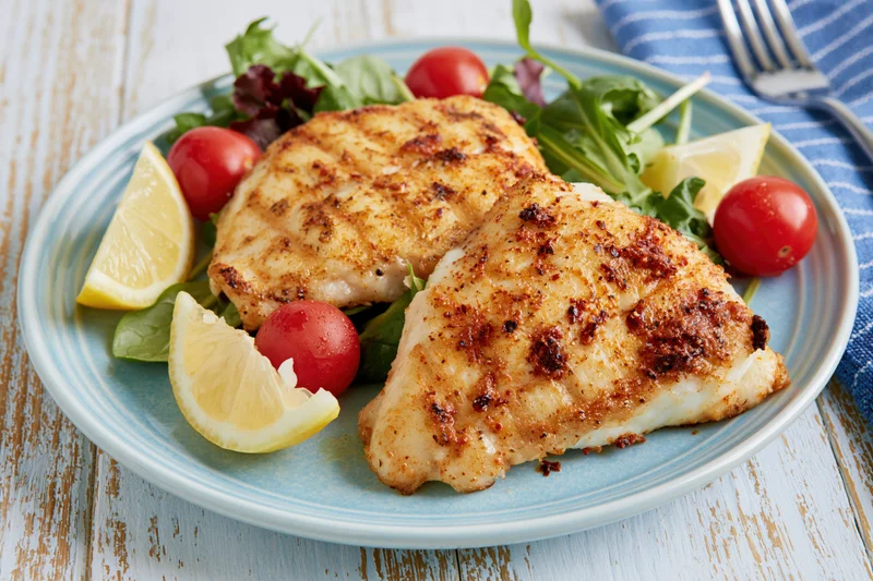 Two seared white fish fillets with lemon and salad on a blue plate