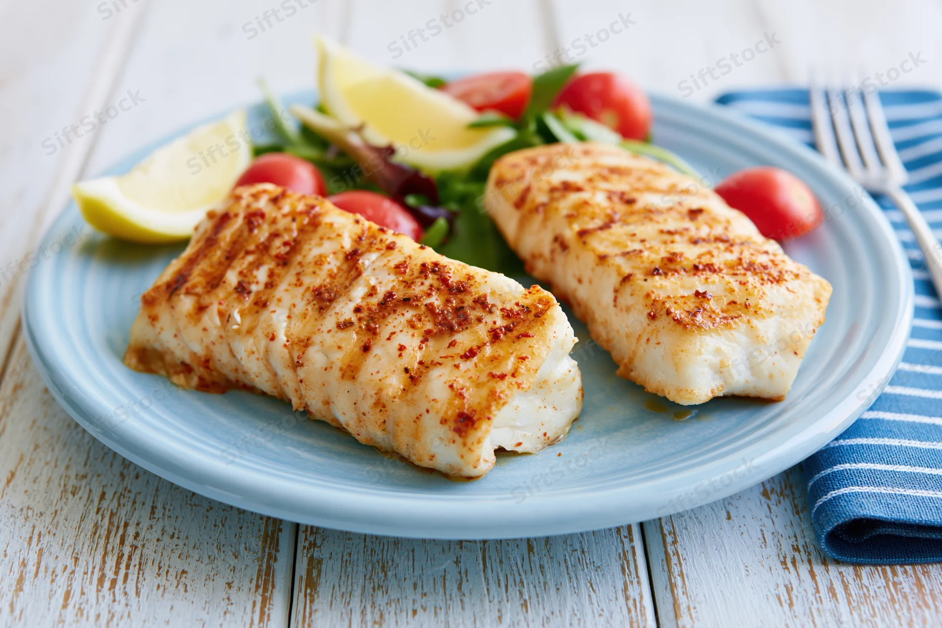 Two grilled white fish fillets with salad and lemon wedges on a blue plate on a wooden table