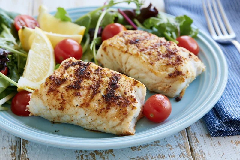 Two seared white fish fillets with salad and lemon on a blue plate on a wooden table