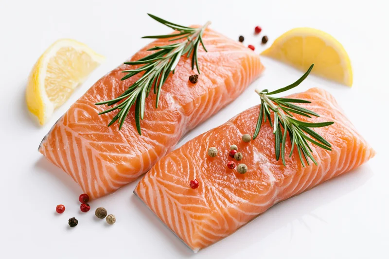 Two raw salmon fillets garnished with rosemary, lemon, and peppercorns on a white surface