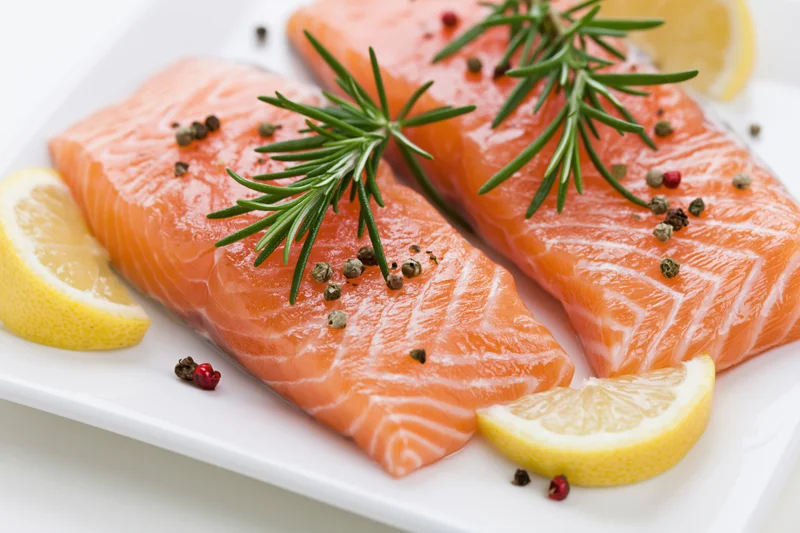 Two raw salmon fillets garnished with rosemary, lemon, and peppercorns on a white plate