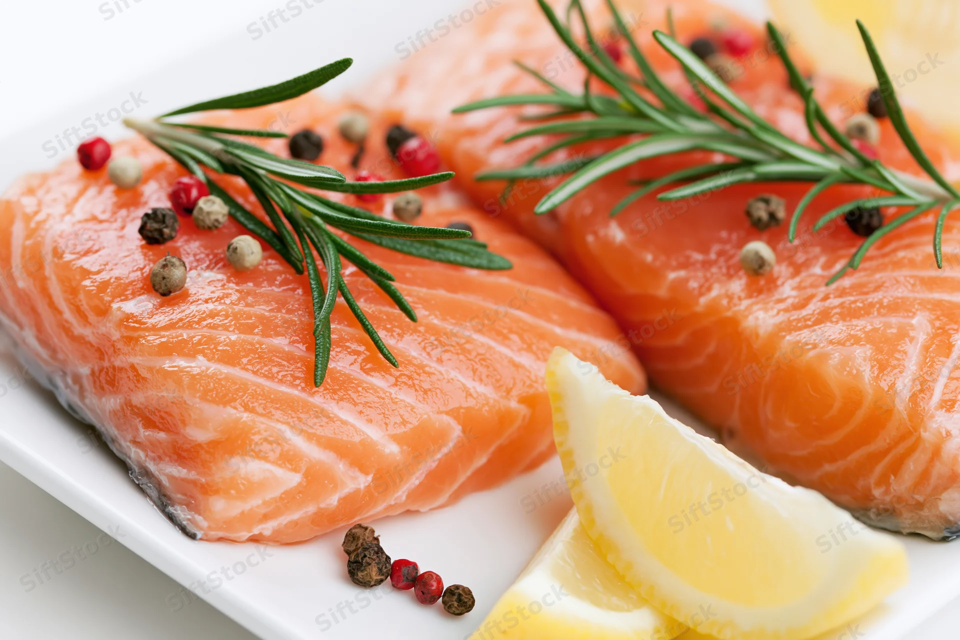 Two raw salmon fillets garnished with rosemary, lemon, and peppercorns on a white plate