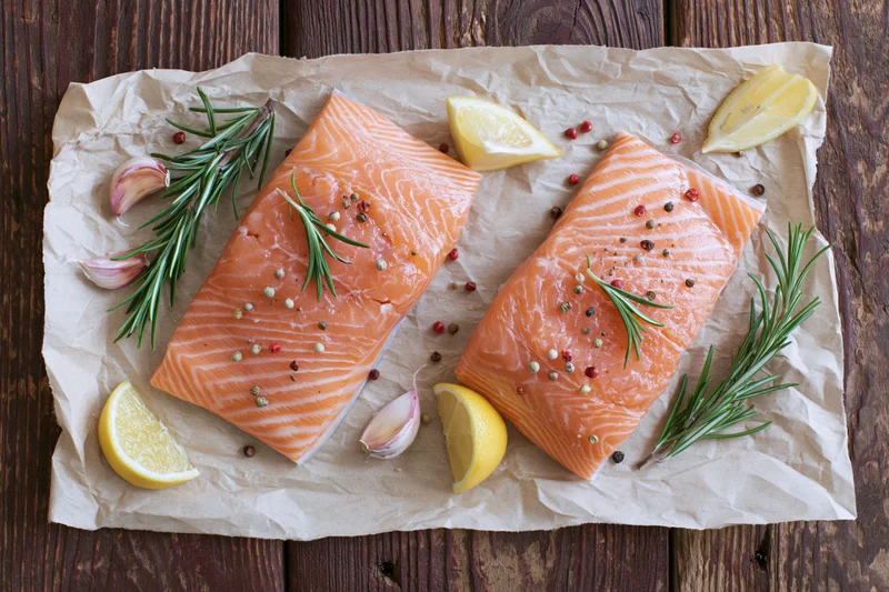 Two raw salmon fillets seasoned with herbs and lemon on crumpled parchment paper over wooden surface