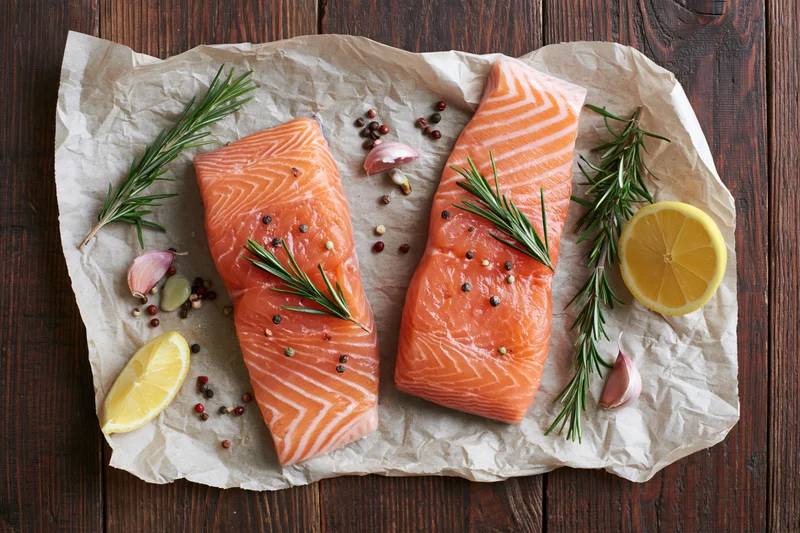 Two raw salmon fillets garnished with rosemary, lemon, and garlic on crumpled parchment paper over wooden table