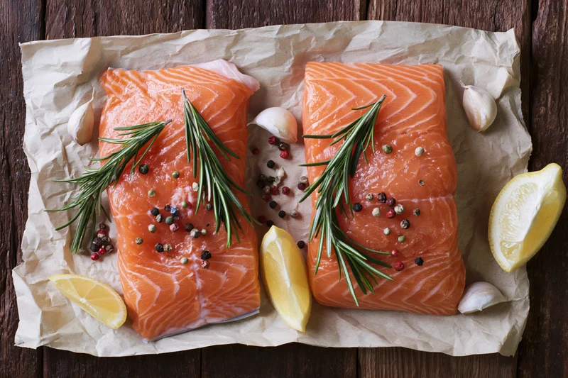Two raw salmon fillets seasoned with rosemary, peppercorns, garlic, and lemon wedges on crumpled parchment paper over wooden surface