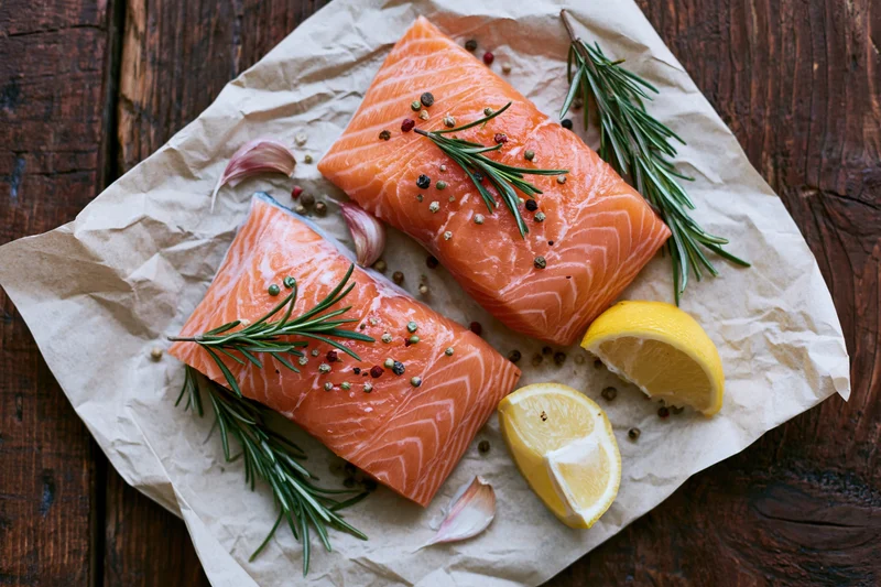 Two raw salmon fillets garnished with rosemary, lemon, and garlic on crumpled parchment paper over wooden table