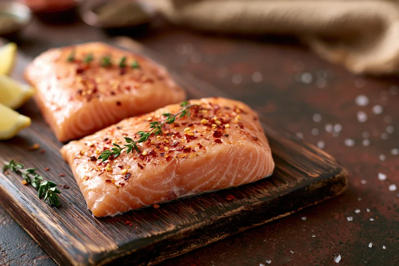Two raw salmon fillets seasoned with chili flakes and thyme on a wooden cutting board