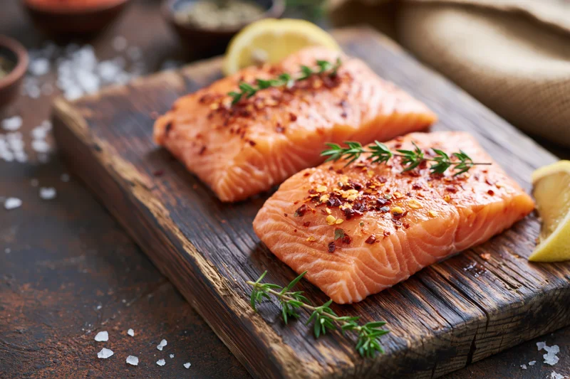 Two raw salmon fillets seasoned with chili flakes and thyme on a wooden cutting board