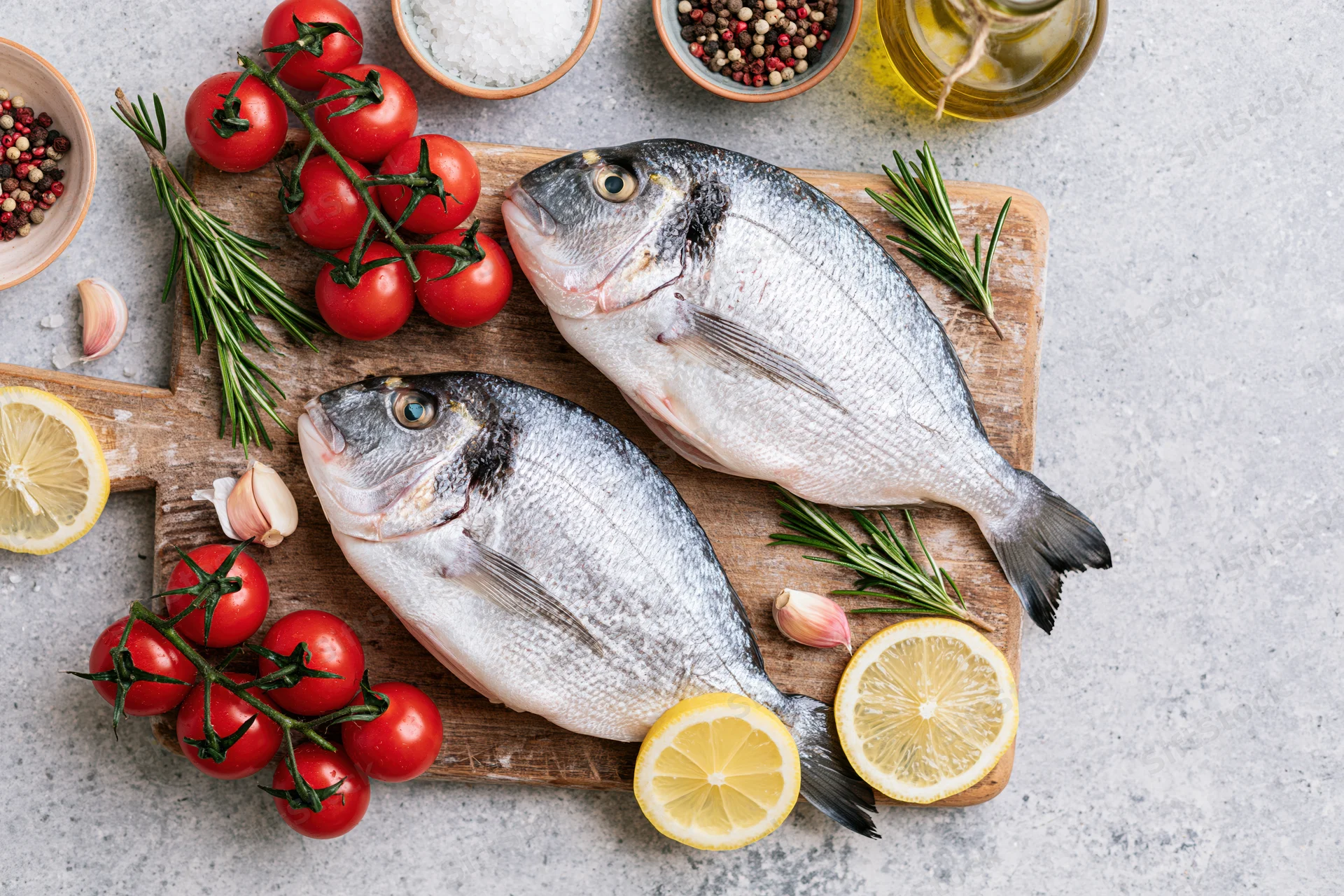 Two raw sea bream fish on a wooden cutting board surrounded by tomatoes, lemon, rosemary, garlic, and spices