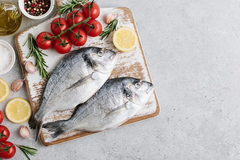 Two raw sea bream fish on a worn wooden cutting board with tomatoes, lemon, herbs and spices around