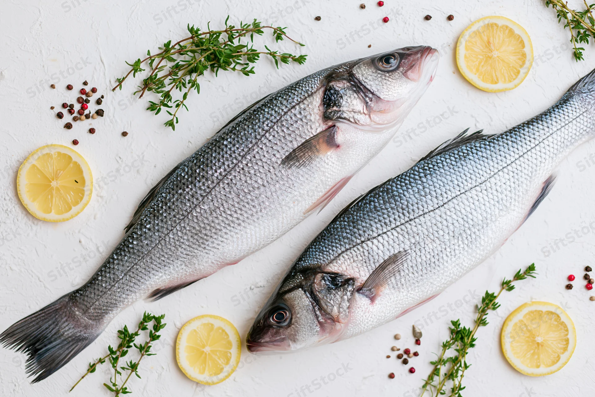 Two raw whole sea bass fish arranged with lemon slices and herbs on a white textured surface