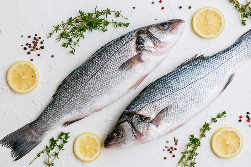 Two raw whole sea bass fish arranged with lemon slices and herbs on a white textured surface