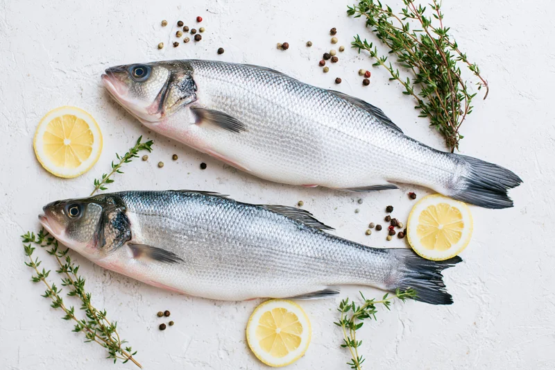 Two raw whole sea bass fish arranged with lemon slices, thyme, and peppercorns on a white textured surface