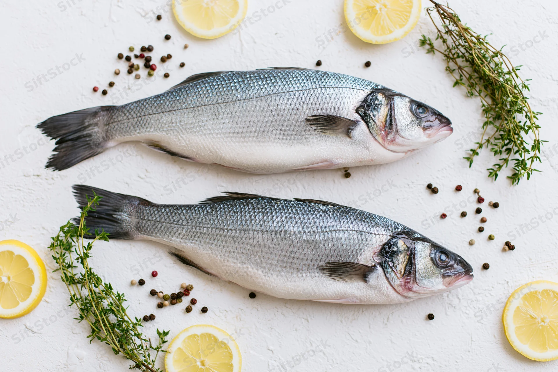 Two raw whole sea bass fish arranged with lemon slices, thyme, and peppercorns on a white textured surface
