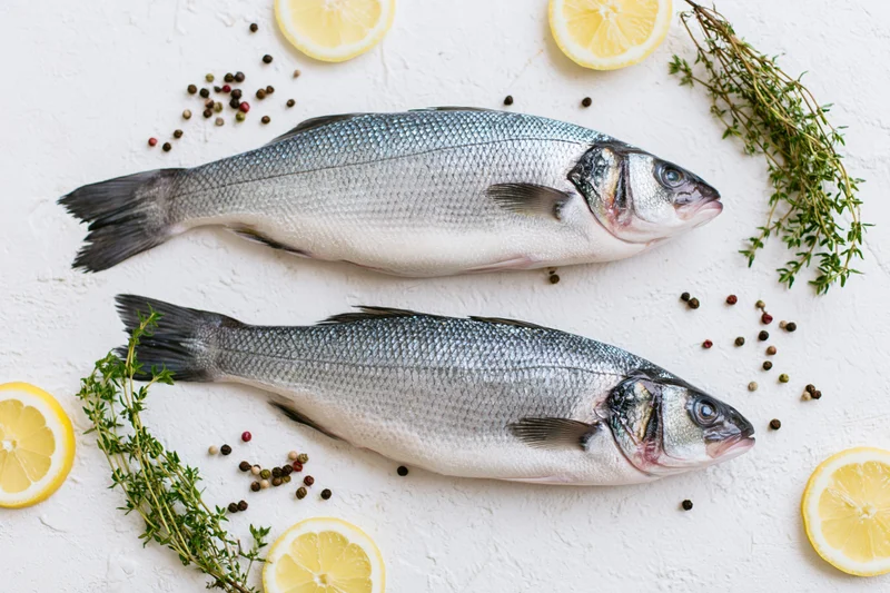Two raw whole sea bass fish arranged with lemon slices, thyme, and peppercorns on a white textured surface