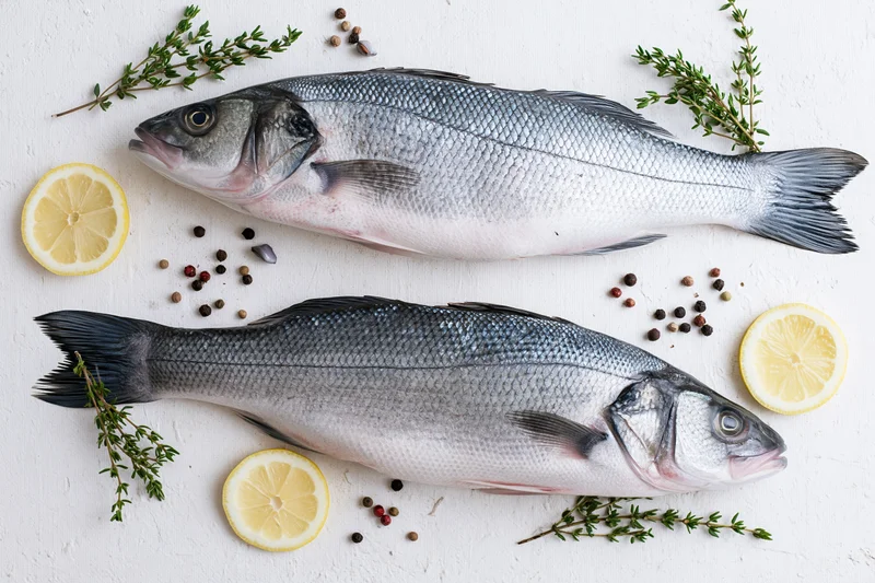 Two raw whole sea bass fish arranged with lemon slices, thyme, and peppercorns on a white textured surface