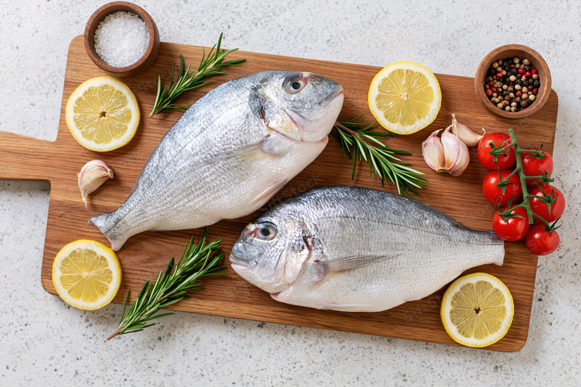 Two raw whole sea bream on wooden cutting board with lemon, rosemary, garlic, tomatoes, salt, pepper