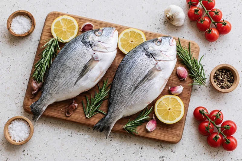 Two raw whole sea bream fish on a wooden cutting board with lemon, rosemary, garlic, cherry tomatoes, salt, and pepper arranged around them on a white speckled countertop